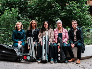 Fem musikere sitter på en mur foran en grønn busk.